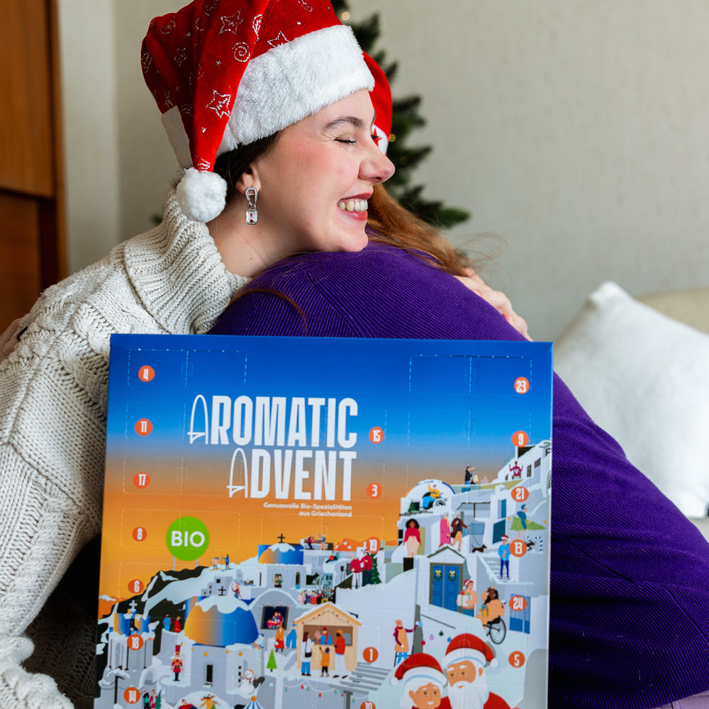 Aroma Olymp. Frau mit Weihnachtsmütze hält den Bio Feinkost Adventskalender 2025 und umarmt eine Person.