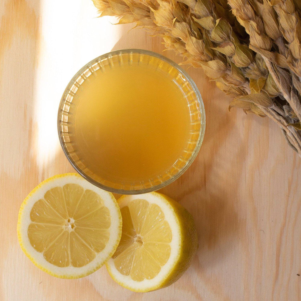 Ein Glas, gefüllt mit Aroma Olymp Craft Tea Still, steht auf einem Tisch. Daneben liegt eine halbierte Zitrone und einige Zweige Griechischer Bergtee.